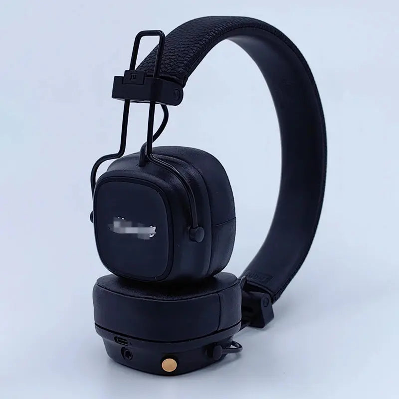 Black over-ear headphones with a padded headband, cushioned ear cups, and a small gold circular detail on the lower side, photographed on a clean light gray studio background.